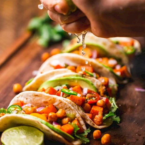 A hand squeezing lime over a row of tacos filled with chickpeas, tomato, onion, avocado, and cilantro, on a wooden board.