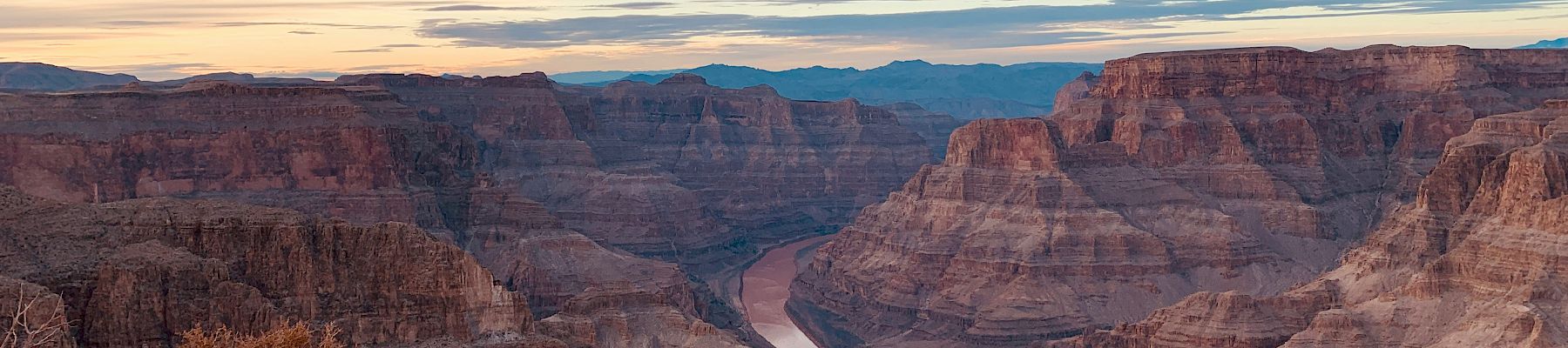 A vast canyon with layered rock formations and a winding river at the bottom, under a sky filled with wispy clouds and a hint of sunset.