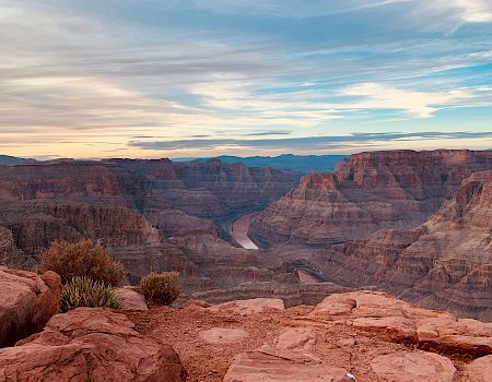 A vast canyon with layered rock formations and a winding river at the bottom, under a sky filled with wispy clouds and a hint of sunset.